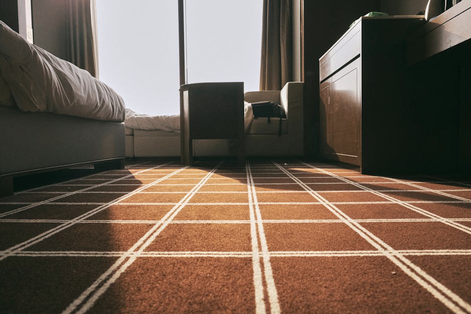 A low-angle view of a hotel or bedroom interior during daylight, featuring a checkered brown carpet with white grid lines, a bed with white bedding on the left, a small wooden dresser or cabinet on th
