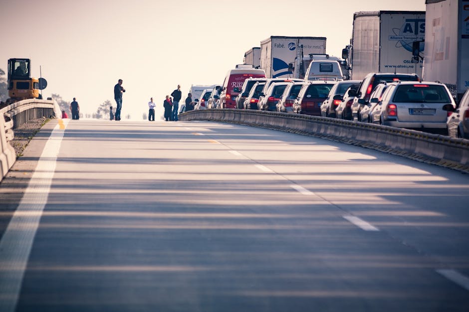 Image showing a highway or service road with multiple parked vehicles, including trucks and passenger cars, lined up along the shoulder. The road surface appears clean and well-maintained, with visibl