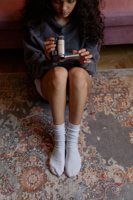 A close-up view of a modern vacuum cleaner with a black handle and a wide, flat cleaning head, actively cleaning a patterned area rug with intricate floral designs in muted tones. The rug appears clea